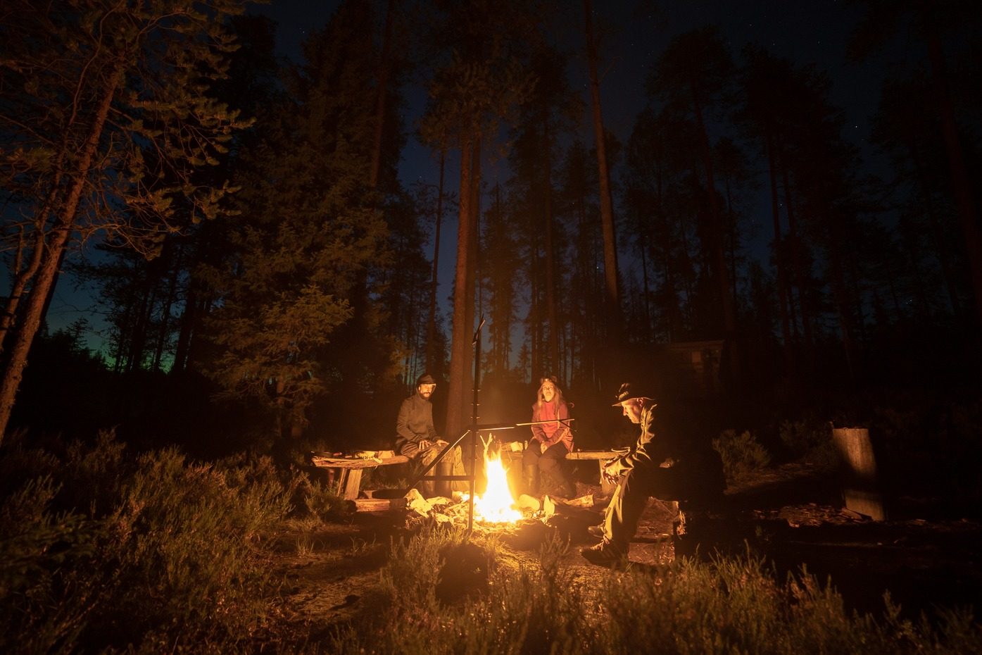 Taruma tribe at the campfire in the Kuhmo night — three people talking by the fire under starry sky and northern lights.
